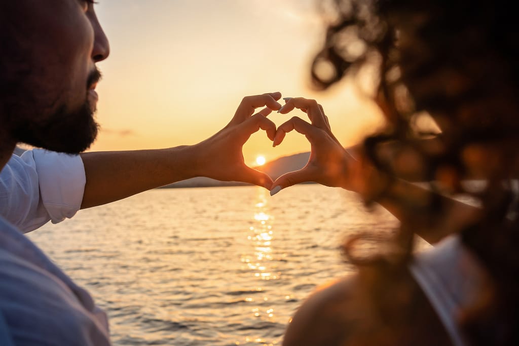 Couple making a heart with their hands over the water during the sun setting.