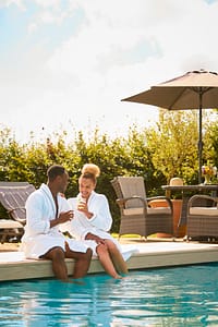 Couple Wearing Robes Outdoors Sitting With Drinks With Feet In Swimming Pool On Spa Day Couple enjoying drinks by the pool on their honeymoon or anniversary trip