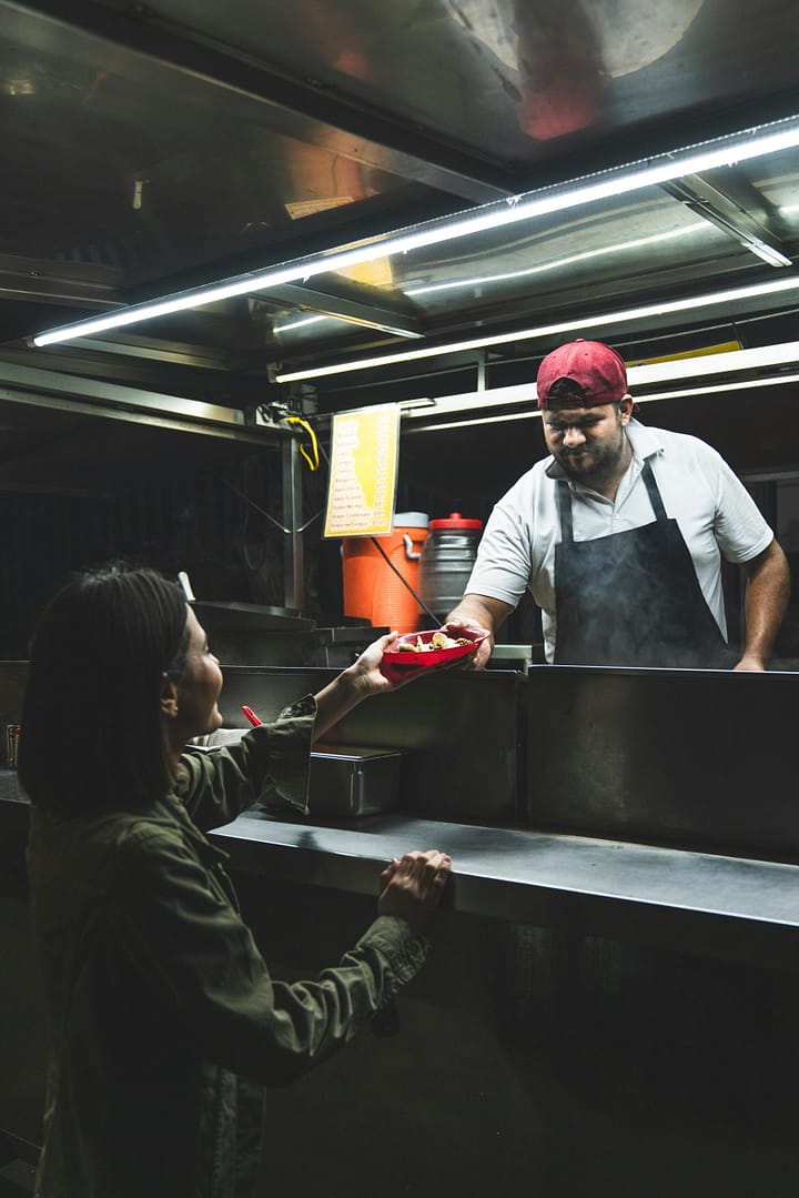 pexels-cristian-rojas-8448070 Woman receives her taco takeout food from male vendor.